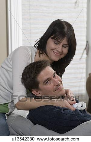 Stock Image - Woman holding compact flourescent lightbulb leans over man seated on sofa, Winnipeg, Canada. Fotosearch