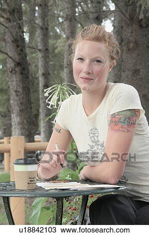 Stock Image - Woman seated at outdoor restaurant, corn-based compostable coffee cup on table, Wasagaming, Riding Mountain National Park, Manitoba, Canada. Fotosearch