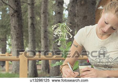 Stock Photography - Woman seated at outdoor restaurant holding corn-based compostable coffee cup, writing, Wasagaming, Riding Mountain National Park, Manitoba, Canada. Fotosearch