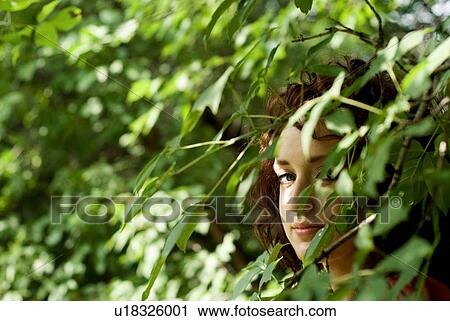 Stock Image - Young woman peeks from behind green leaves, Munson Park, Winnipeg, Canada. Fotosearch