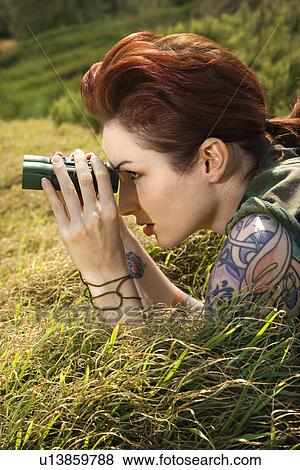 Stock Photo - Attractive tattooed Caucasian woman lying on grass looking through binoculars . Fotosearch