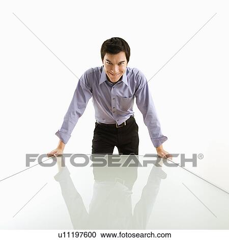 Stock Image - Young Asian business man standing over conference table with devilish grin.. Fotosearch