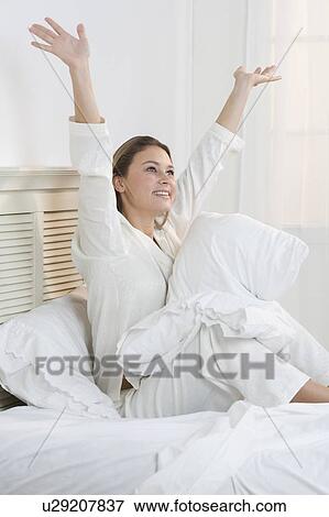 Stock Photo - Woman sitting on bed with her arms raised. Fotosearch