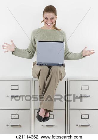 Stock Image - Woman with laptop atop file cabinets. Fotosearch