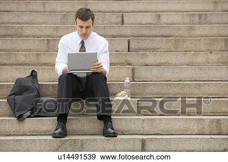 Stock Photo - Young adult male sitting outdoors on stairs with laptop. Fotosearch