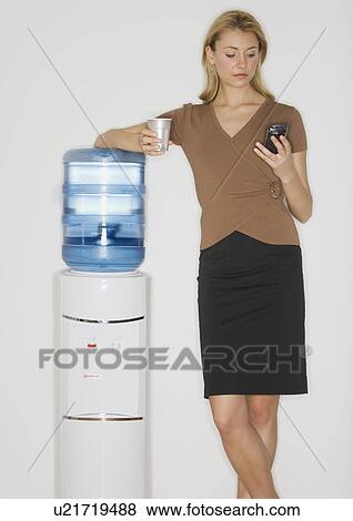 Businesswoman looking at cell phone next to water cooler View Large Photo Image Stock Photo - Businesswoman looking at cell phone next to water cooler. Fotosearch