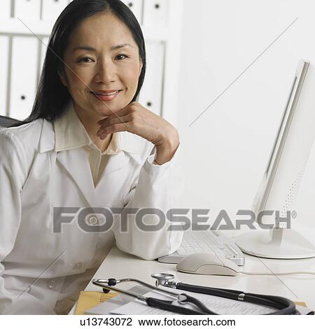 Stock Image - Female doctor sitting at computer smiling. Fotosearch