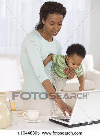Stock Photography - Mother holding baby and typing on laptop. Fotosearch