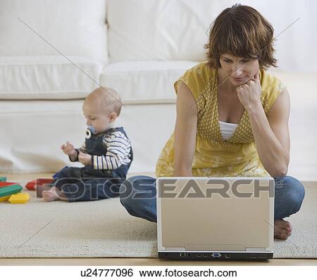 Stock Photograph - Mother using laptop while baby plays. Fotosearch