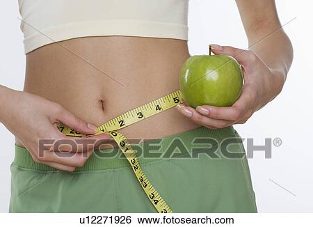Stock Photograph - Studio shot of woman holding apple and measuring her waist. Fotosearch