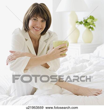 Stock Photo - Woman sitting on bed holding coffee. Fotosearch