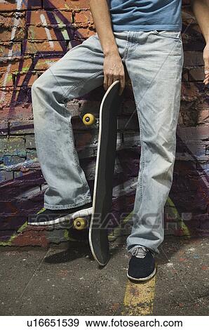 Asian man holding skateboard in front of graffitied wall View Large Photo Image Stock Photo - Asian man holding skateboard in front of graffitied wall. Fotosearch