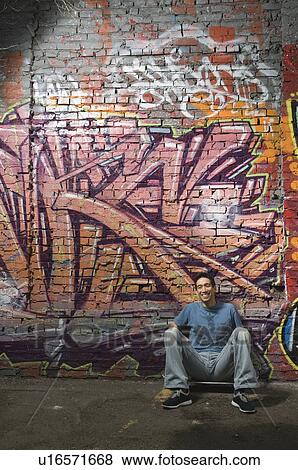 Stock Photo - Asian man sitting in front of graffitied wall. Fotosearch