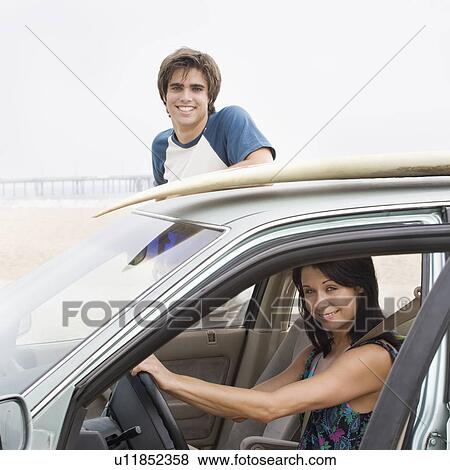 Stock Photo - Couple and car with surfboard on top at beach. Fotosearch
