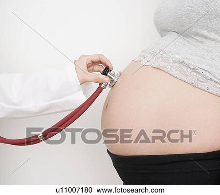 Stock Image - Doctor holding stethoscope on pregnant woman’s belly. Fotosearch
