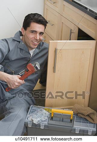 Stock Image - Male plumber holding wrench next to sink. Fotosearch