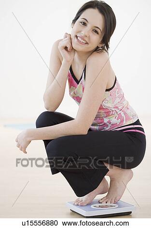 Stock Image - Portrait of woman crouching on bathroom scale. Fotosearch