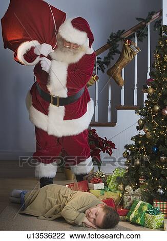 Stock Image - Santa Claus looking at boy sleeping next to Christmas tree. Fotosearch