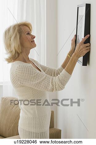 Stock Photograph - Senior woman hanging picture on wall. Fotosearch