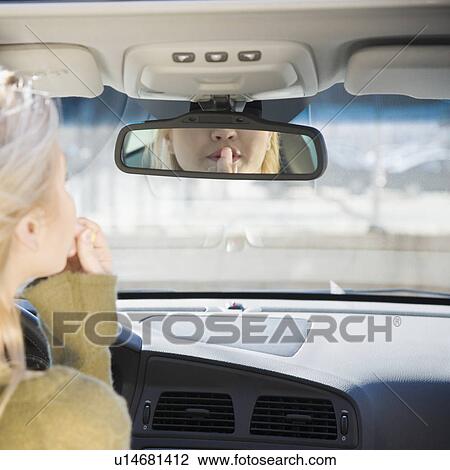 Stock Image - Woman applying lipstick in car rear view mirror. Fotosearch