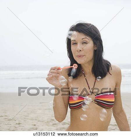 Stock Image - Woman in bikini blowing bubbles at beach. Fotosearch