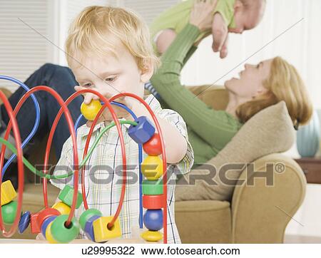 Stock Image - Young son playing with block toy with mother and baby in background. Fotosearch