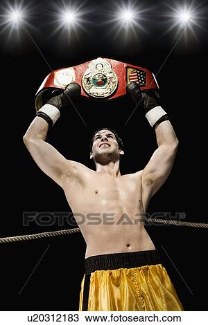 Stock Image - Boxer holding championship belt overhead in boxing ring. Fotosearch