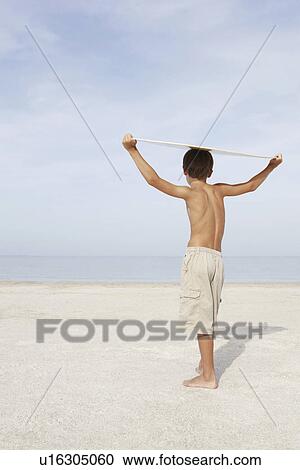 Stock Image - Boy standing on beach holding bodyboard overhead. Fotosearch