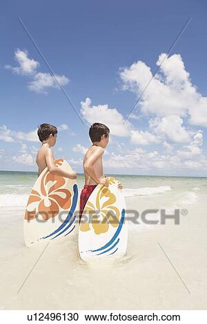 Stock Image - Boys holding skimboards in ocean. Fotosearch