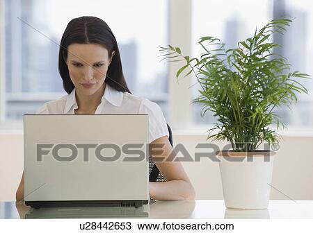 Stock Image - Businesswoman working on laptop next to plant. Fotosearch