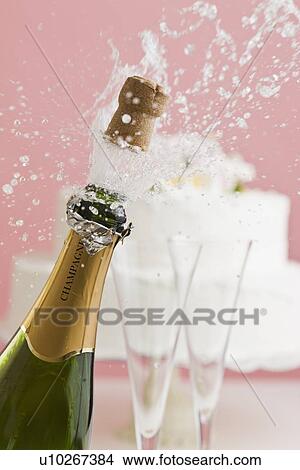Picture - Cork exploding from champagne bottle with wedding cake in background. Fotosearch
