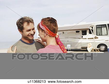 Stock Image - Couple hugging by motor home on beach. Fotosearch