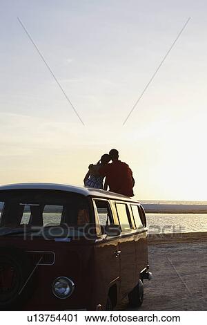 Stock Image - Couple watching ocean sunset from top of van. Fotosearch