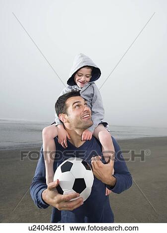 Stock Image - Father carrying son on shoulders and holding soccer ball on beach. Fotosearch