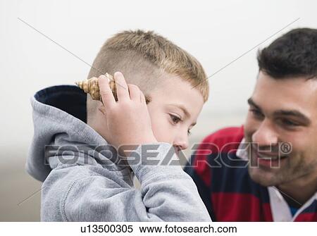 Stock Photography - Father watching son hold seashell up to ear. Fotosearch