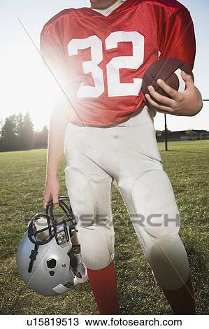Stock Image - Football player holding helmet and football on field. Fotosearch