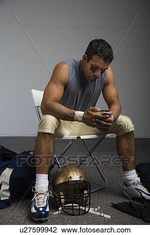 Stock Image - Football player looking at cell phone in locker room. Fotosearch