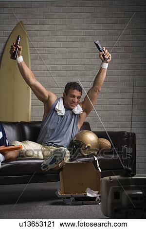 Stock Image - Football player watching football and cheering on sofa. Fotosearch