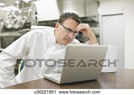 Stock Image - Frustrated chef looking at laptop in kitchen. Fotosearch