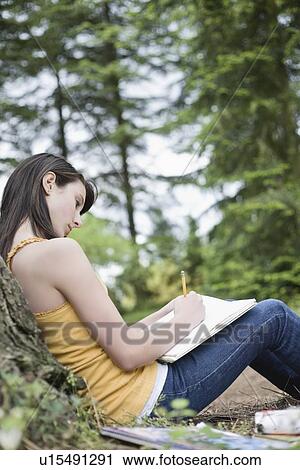 Stock Image - Girl leaning against tree writing in notebook. Fotosearch