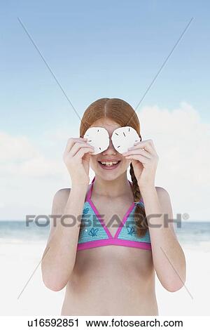 Stock Image - Girl on beach holding sand dollars over eyes. Fotosearch
