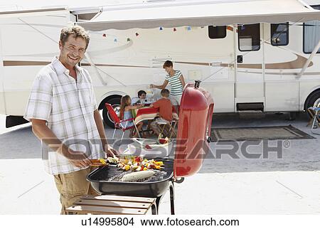 Picture - Man barbecuing with family and motor home in background. Fotosearch
