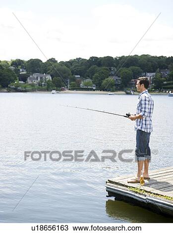 Stock Image - Man fishing off dock with beer at feet. Fotosearch