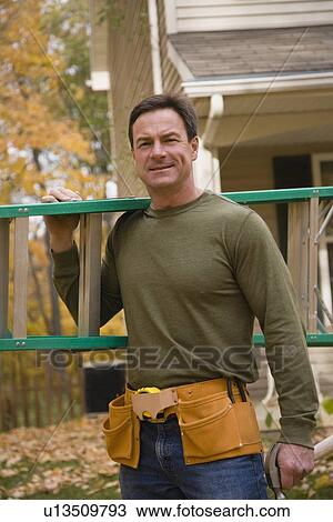 Stock Image - Man holding ladder next to house. Fotosearch