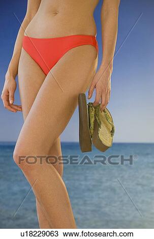 Stock Image - Midsection of woman in bikini holding flip-flops on beach. Fotosearch
