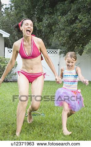 Stock Image - Mother and daughter running through sprinkler. Fotosearch