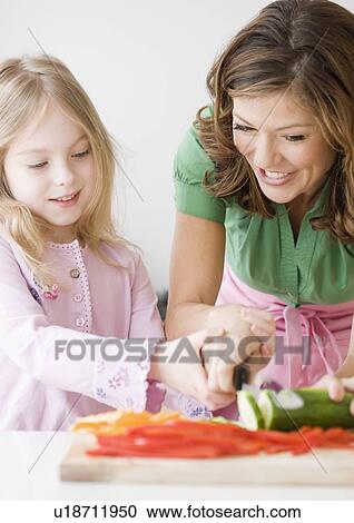 Stock Image - Mother helping daughter chop vegetables. Fotosearch