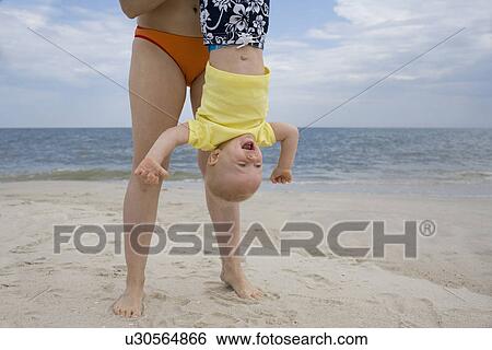 Stock Photograph - Mother holding baby boy upside-down on beach. Fotosearch