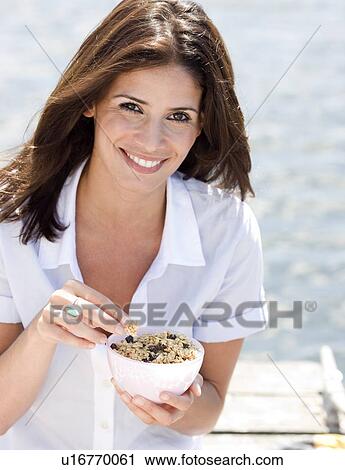 Stock Image - Portrait of woman eating granola on dock. Fotosearch
