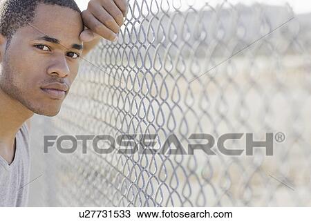 Stock Image - Portrait of young man leaning against chain-link fence. Fotosearch